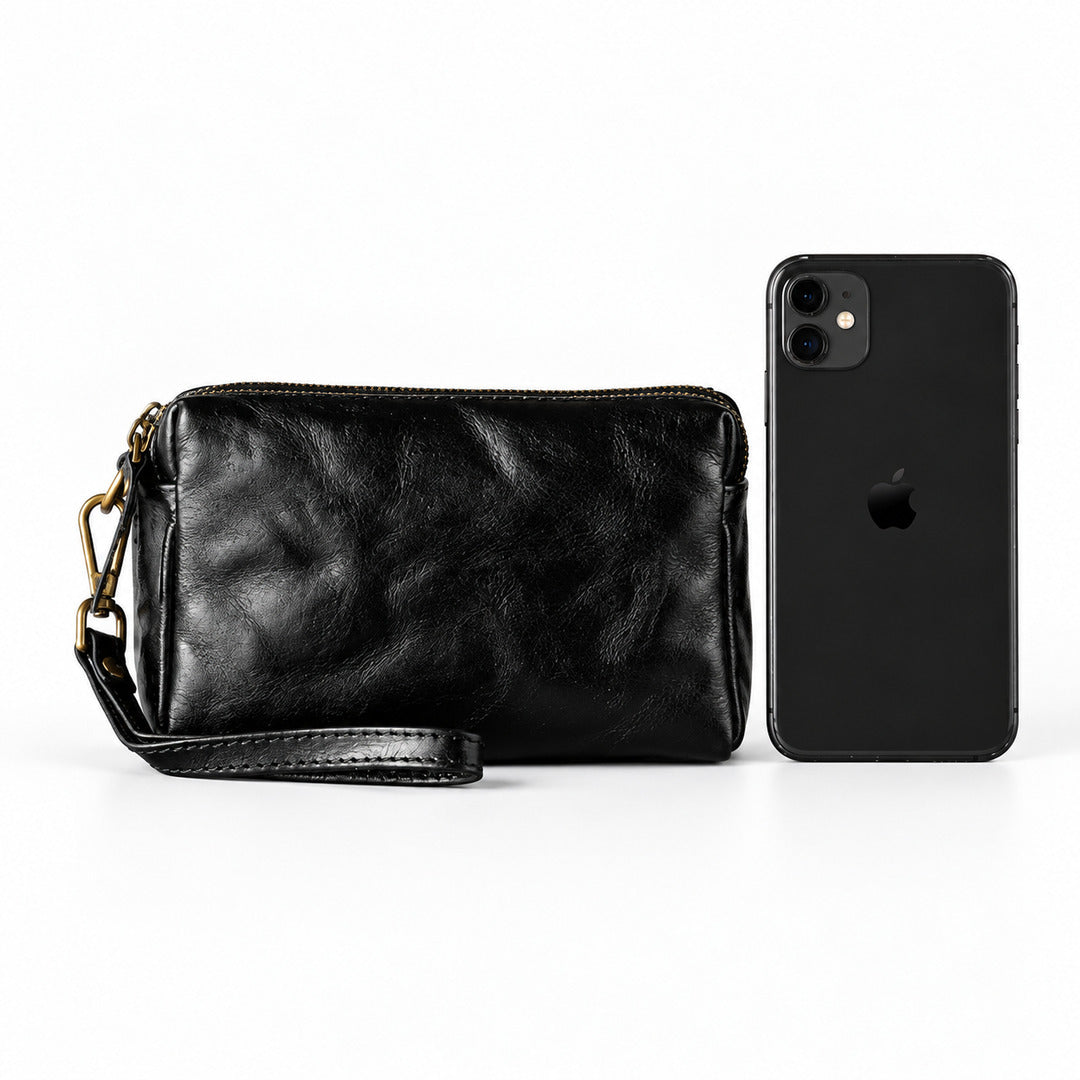 Black leather wristlet clutch with gold-tone hardware and a wrist strap, shown next to a black smartphone.