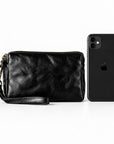 Black leather wristlet clutch with gold-tone hardware and a wrist strap, shown next to a black smartphone.