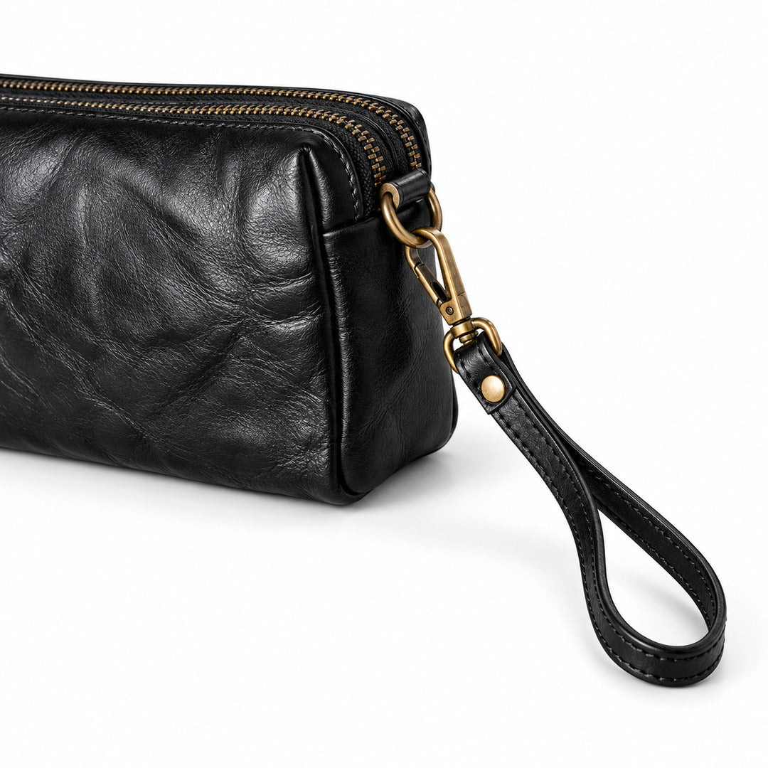 A close-up of a black leather wristlet clutch with a top zipper, gold-tone hardware, and a matching black leather wrist strap.