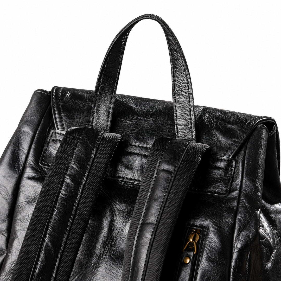 Close-up of a black textured leather backpack showing the top handle, front flap, dual shoulder straps, and a gold-tone zipper pull on a white background.