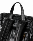 Close-up of a black textured leather backpack showing the top handle, front flap, dual shoulder straps, and a gold-tone zipper pull on a white background.