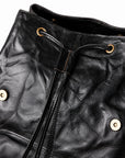 Close-up of a black leather bag with a drawstring closure, brass eyelets, and gold-tone snap buttons on a white background.