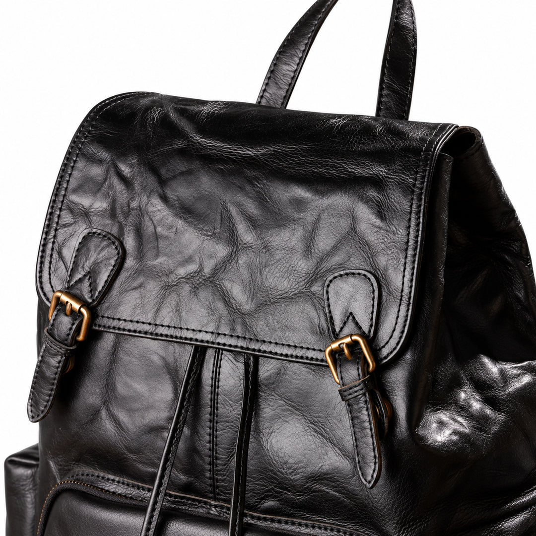 Close-up of a black leather backpack with a wrinkled texture, gold-tone buckle straps on the front flap, and top handles, shown against a white background.