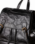 Close-up of a black leather backpack with a wrinkled texture, gold-tone buckle straps on the front flap, and top handles, shown against a white background.