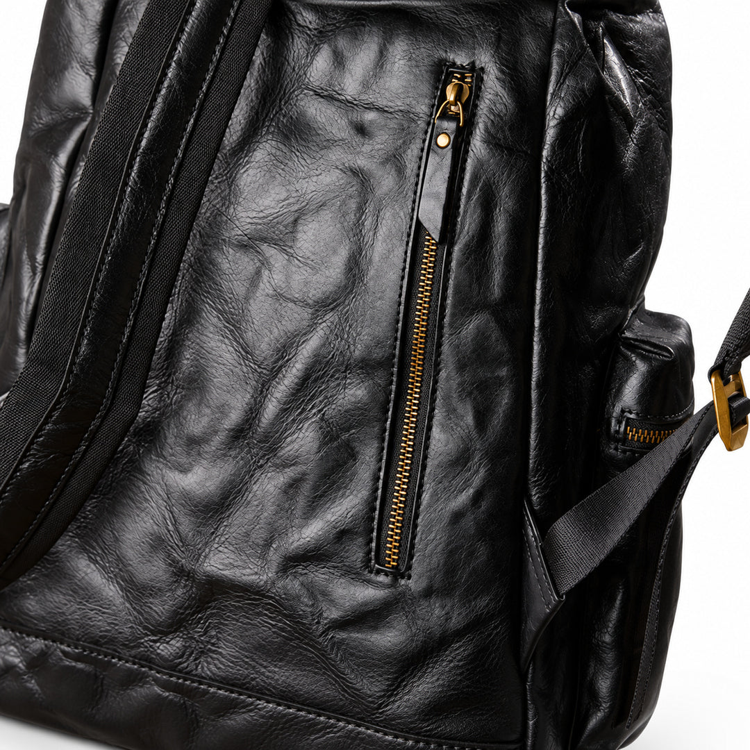 Close-up of a black leather backpack showing the front pocket with a vertical gold-tone zipper, textured leather surface, and part of the side strap on a white background.