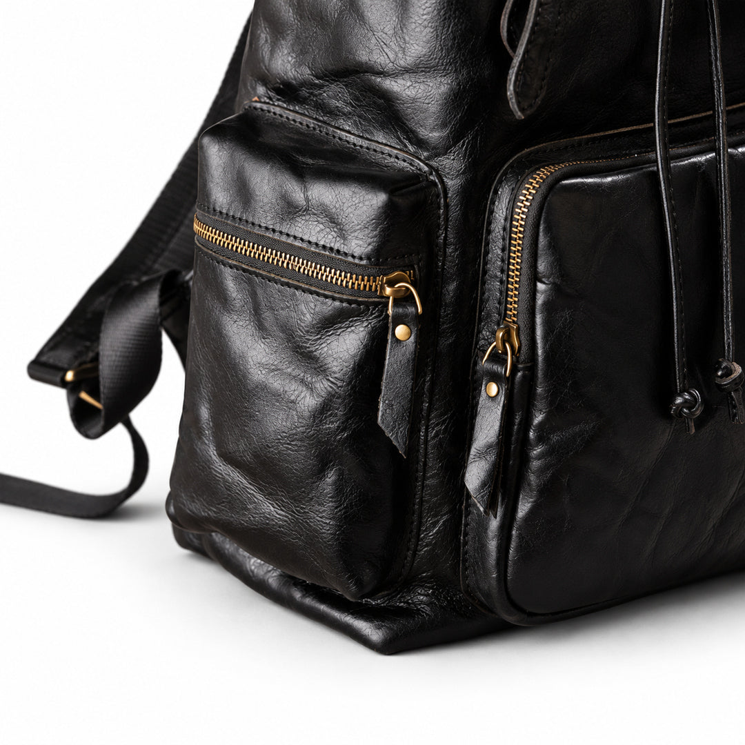 Close-up of a black leather backpack showing the front pockets, gold-tone zippers, and textured leather surface on a white background.