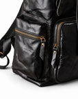 Close-up of a black leather backpack showing the front pockets, gold-tone zippers, and textured leather surface on a white background.