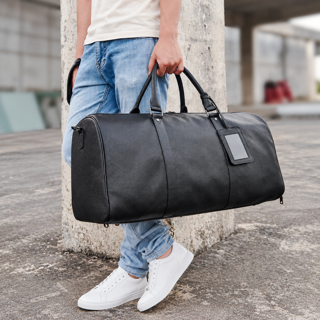 A person outdoors holding a large black leather duffel bag with dual top handles and a blank rectangular patch on the side. The person is wearing light blue jeans and white sneakers, standing beside a concrete pillar.