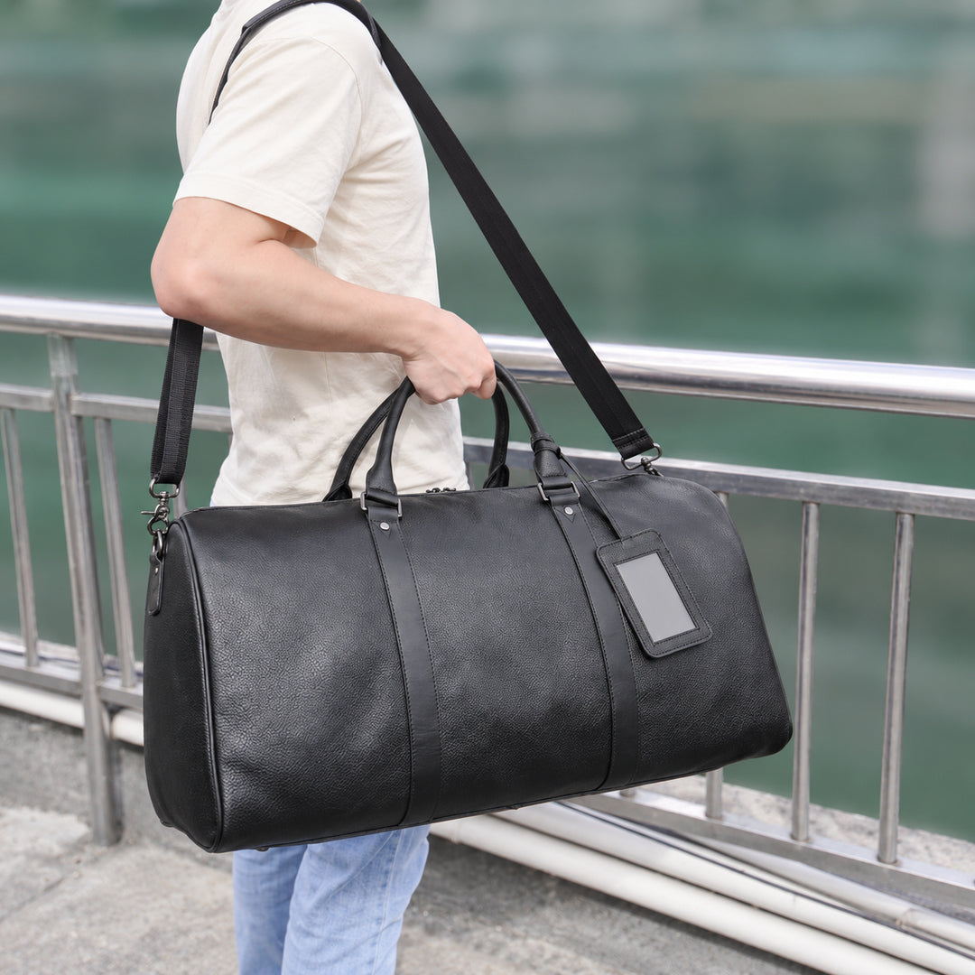A man in a cream t-shirt and light blue jeans carries a large black leather duffel bag with a shoulder strap and a blank luggage tag, standing next to a metal railing with a blurred green background.