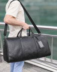 A man in a cream t-shirt and light blue jeans carries a large black leather duffel bag with a shoulder strap and a blank luggage tag, standing next to a metal railing with a blurred green background.