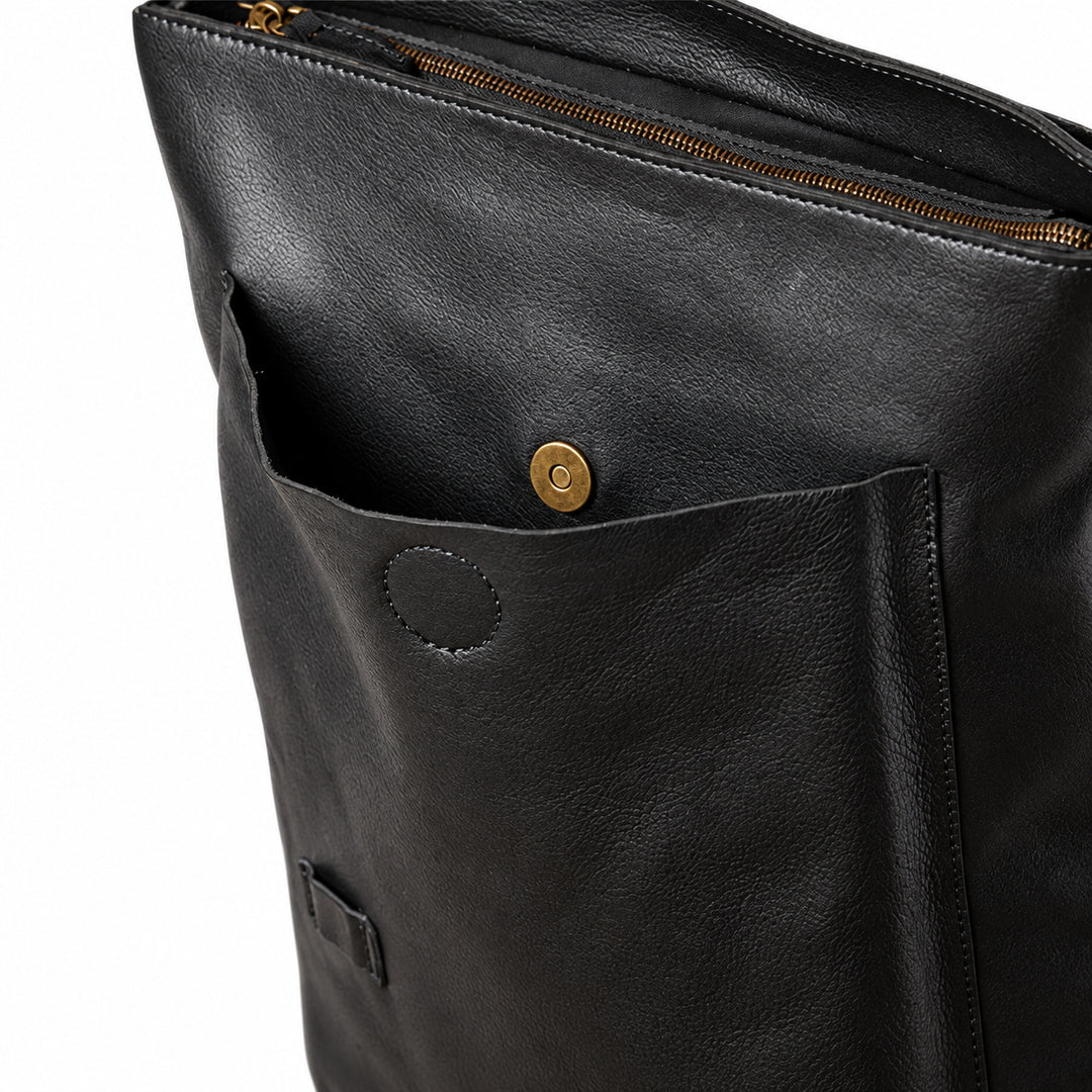 Close-up of a black leather bag showing a front pocket with a brass snap button, a zipper opening at the top, and a small leather tag near the bottom.