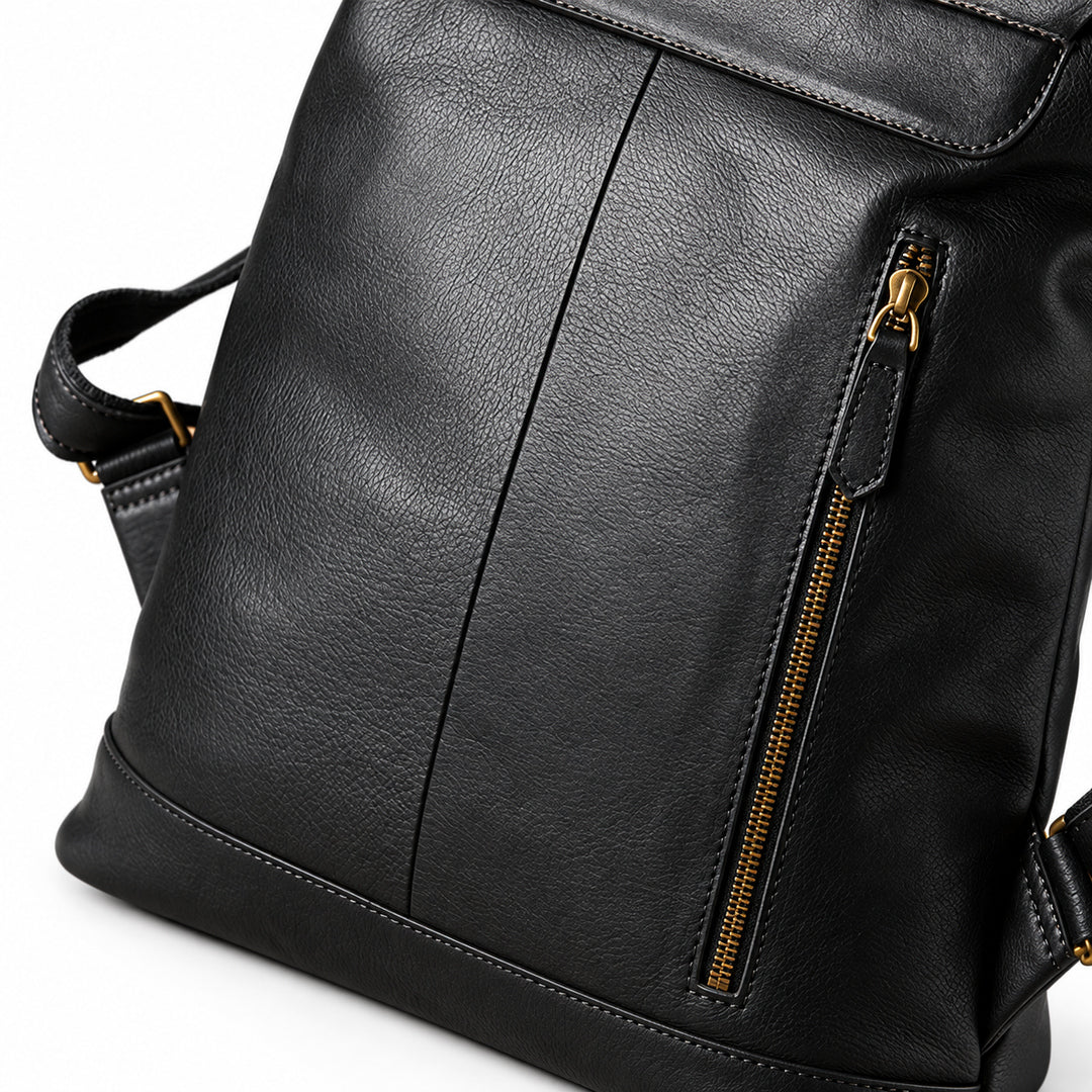 A close-up of a black textured leather backpack or messenger bag with a vertical seam, a side zippered pocket with gold-tone hardware, and a strap visible on the left.
