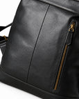 A close-up of a black textured leather backpack or messenger bag with a vertical seam, a side zippered pocket with gold-tone hardware, and a strap visible on the left.