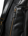 Close-up of a black leather bag showing a vertical zippered pocket with a gold-tone zipper pull and a matching gold buckle on the shoulder strap.