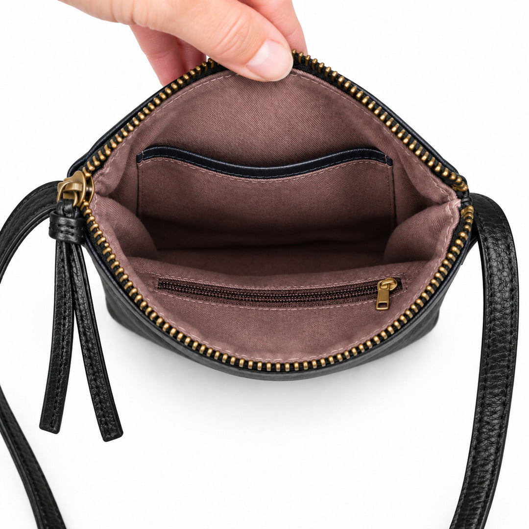 A close-up of a black leather crossbody bag being held open by a hand, showing a brown fabric interior with gold-tone zippers and a small zip pocket.