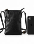 Black leather crossbody phone bag with a top zipper, shown next to a black smartphone for scale on a white background.