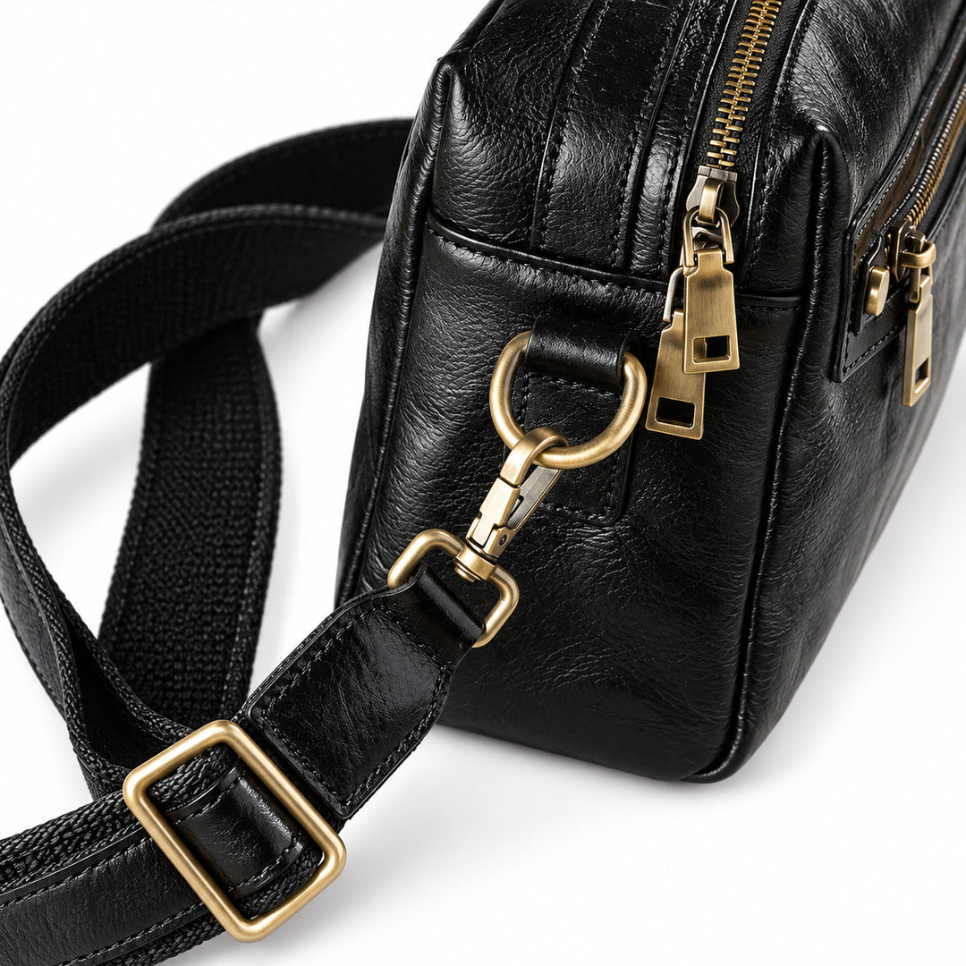 Close-up of a black leather crossbody bag with a wide black webbing strap and gold-tone hardware, including zippers and a ring clasp.