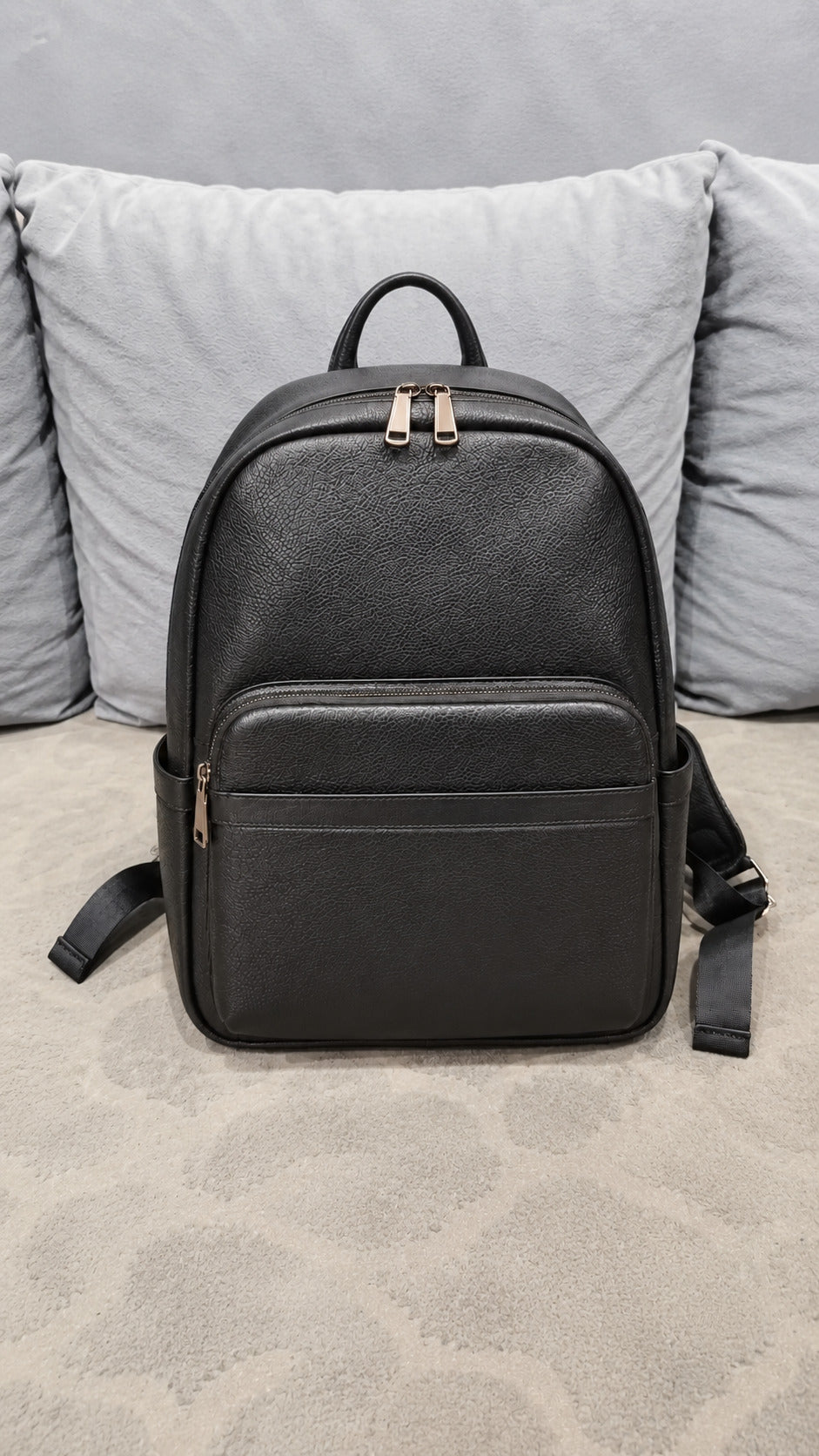 A black textured leather backpack with gold-tone zipper pulls, displayed on a patterned beige carpet in front of gray pillows.