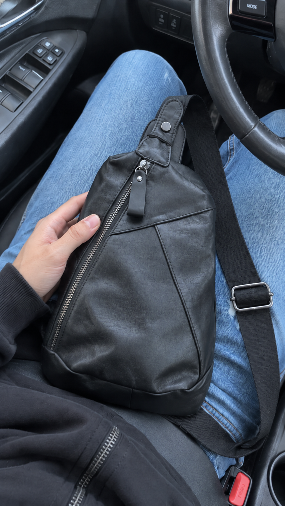 Black leather sling crossbody bag with front zipper and adjustable strap shown inside a car