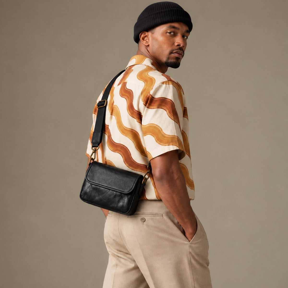 Rear view of a man wearing a black textured crossbody bag at his lower back, with an adjustable strap and brass-tone clips.