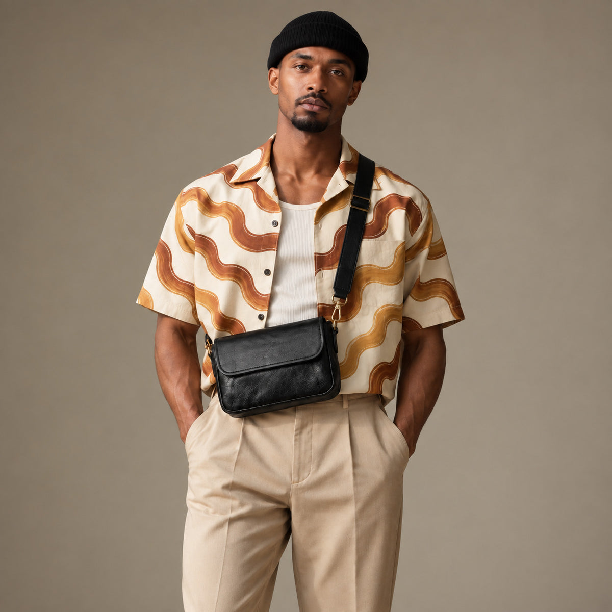 Man wearing a black textured crossbody bag across the front, with a flap closure, wide strap and brass-tone hardware.