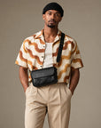 Man wearing a black textured crossbody bag across the front, with a flap closure, wide strap and brass-tone hardware.