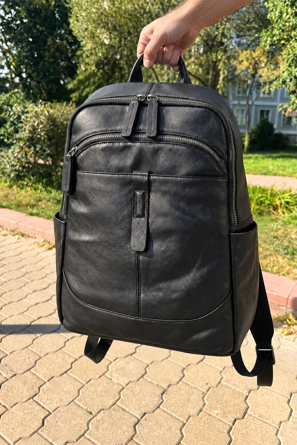 A black leather backpack held by a hand outdoors on a paved walkway with trees and a building in the background.
