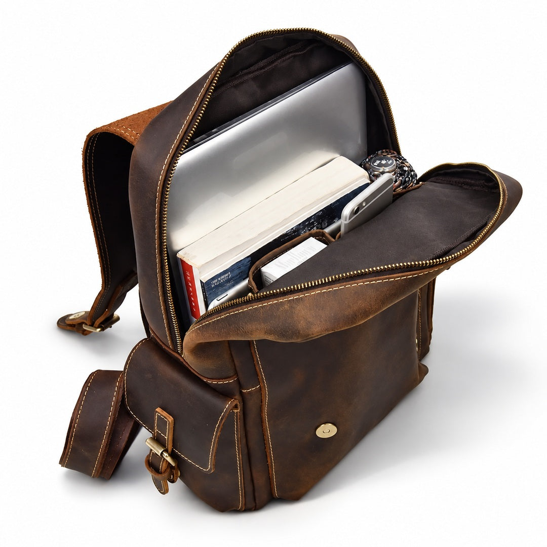 A brown distressed leather crossbody bag shown open, filled with a laptop, books, and a watch.