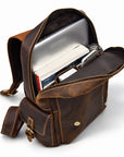 A brown distressed leather crossbody bag shown open, filled with a laptop, books, and a watch.