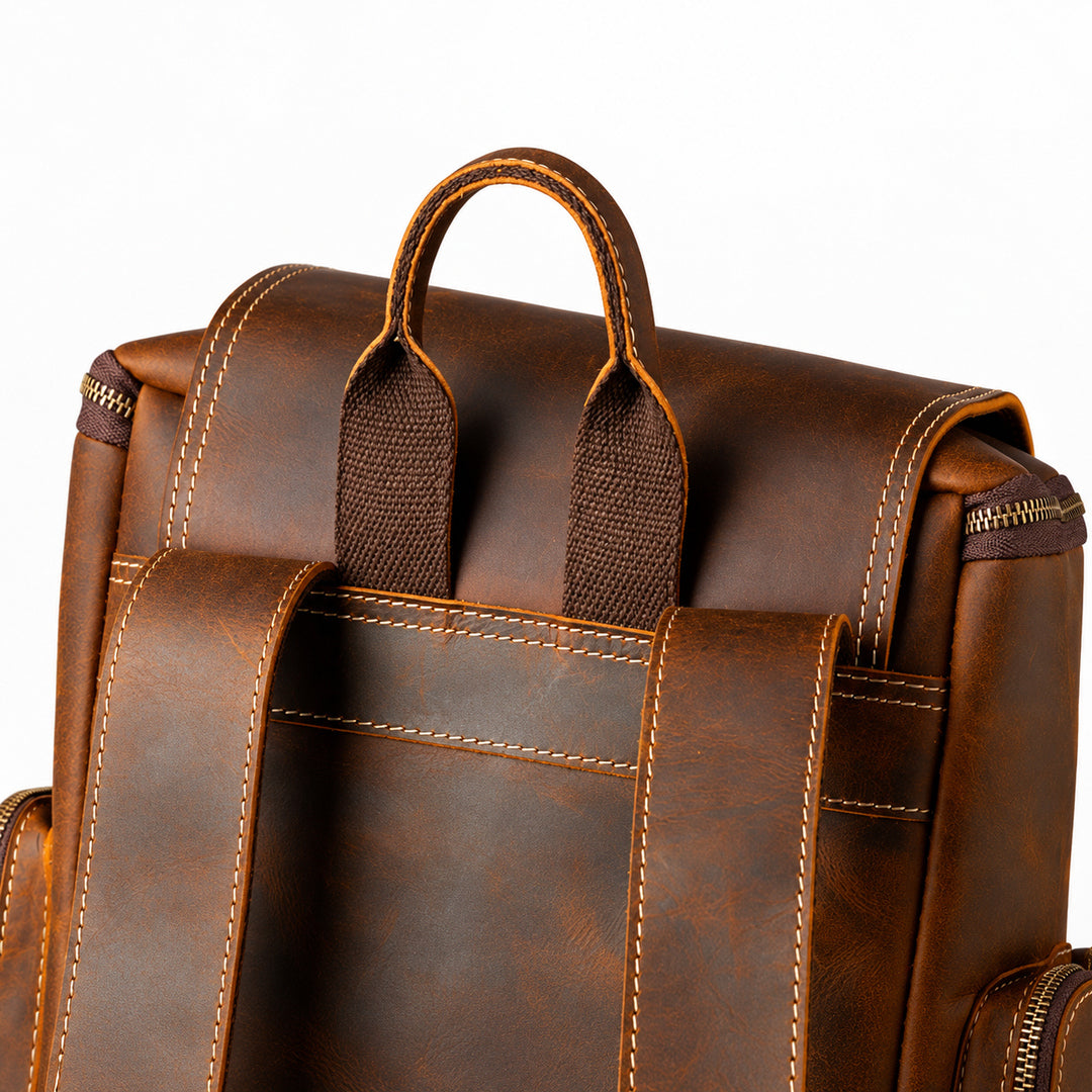 Close-up of the back of a brown leather backpack showing the handle, shoulder straps, zipper, and contrast stitching.