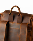 Close-up of the back of a brown leather backpack showing the handle, shoulder straps, zipper, and contrast stitching.