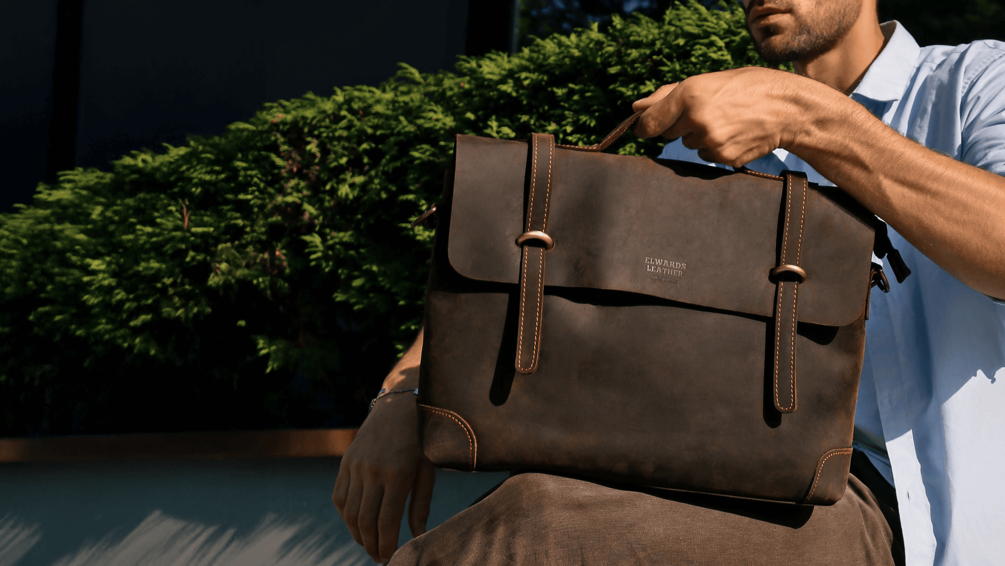 Man holding a brown leather bag outdoors with greenery in the background