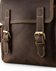 Close-up of a dark brown leather bag with two front buckle straps, white contrast stitching, and brass-tone.