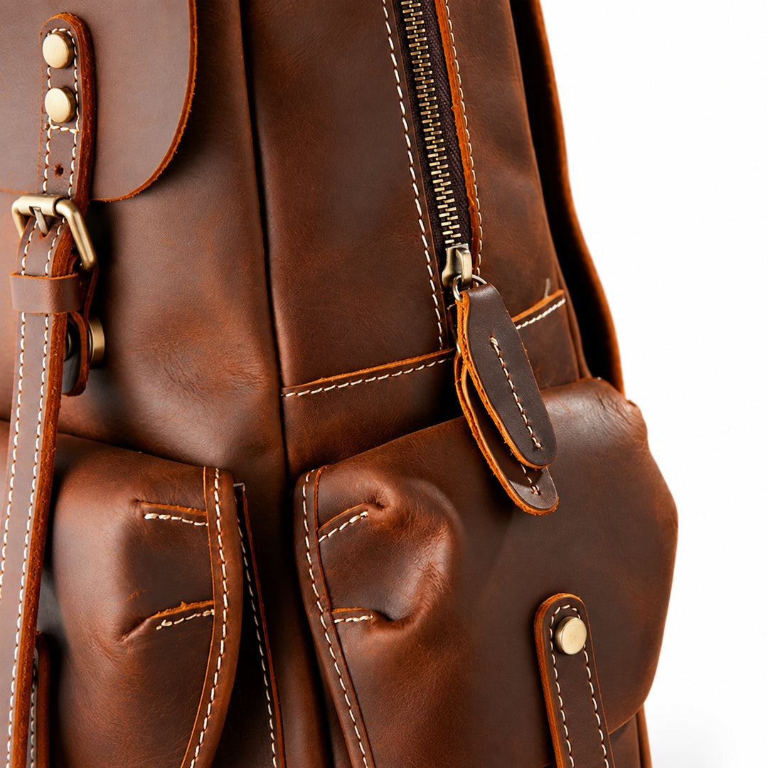 Close-up of a brown leather backpack showing the front pockets, zipper, stitching, and brass-tone hardware.
