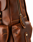 Close-up of a brown leather backpack showing the front pockets, zipper, stitching, and brass-tone hardware.