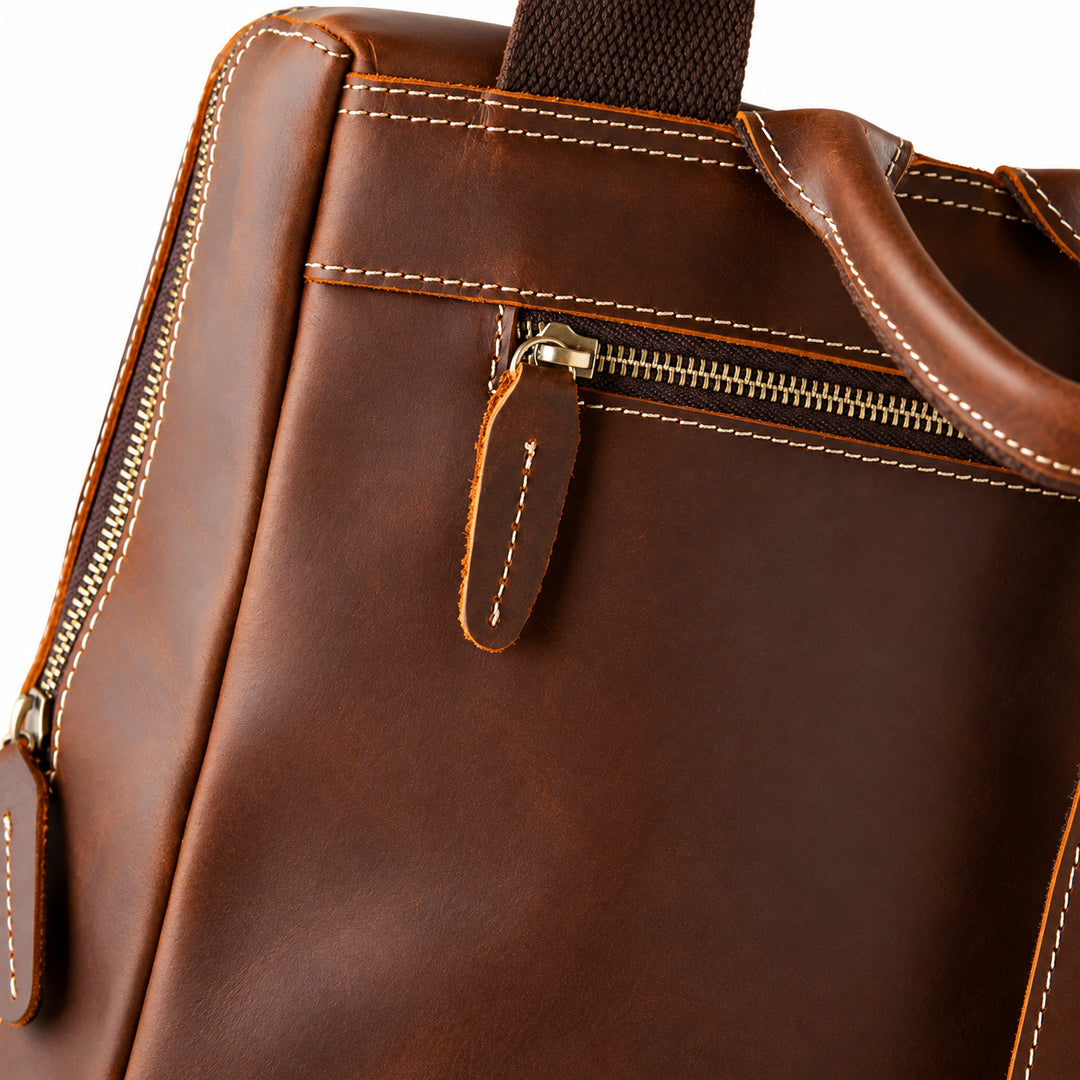 Close-up of a brown leather bag showing a front zipper pocket with white contrast stitching, a leather pull tab, and part of a dark webbed handle.
