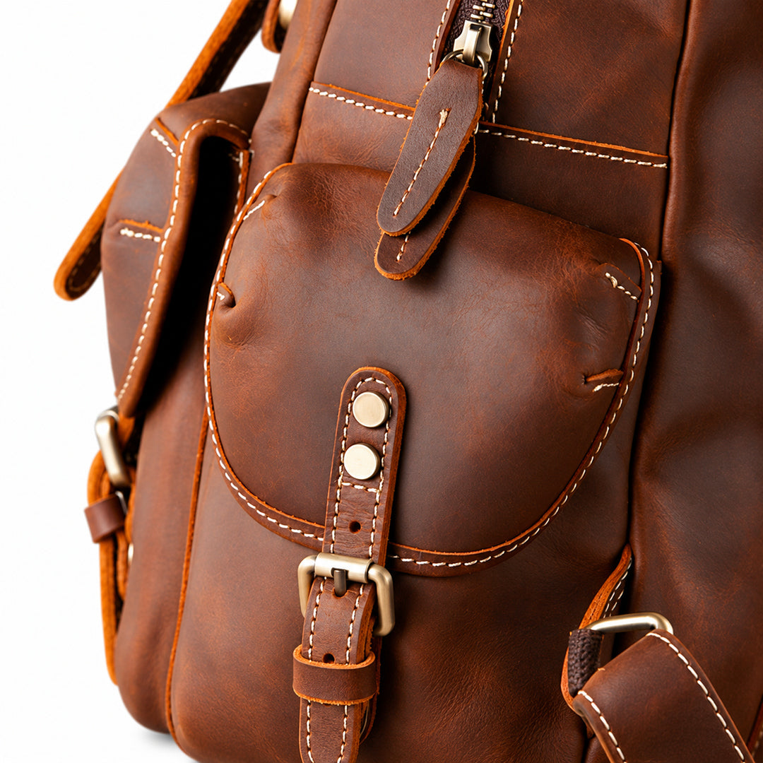 Close-up of a brown leather backpack showing the front pocket, stitching, buckle strap, and metal hardware.