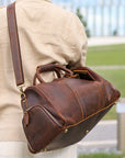 Stylish brown leather weekender bag with adjustable strap shown on shoulder outdoors.