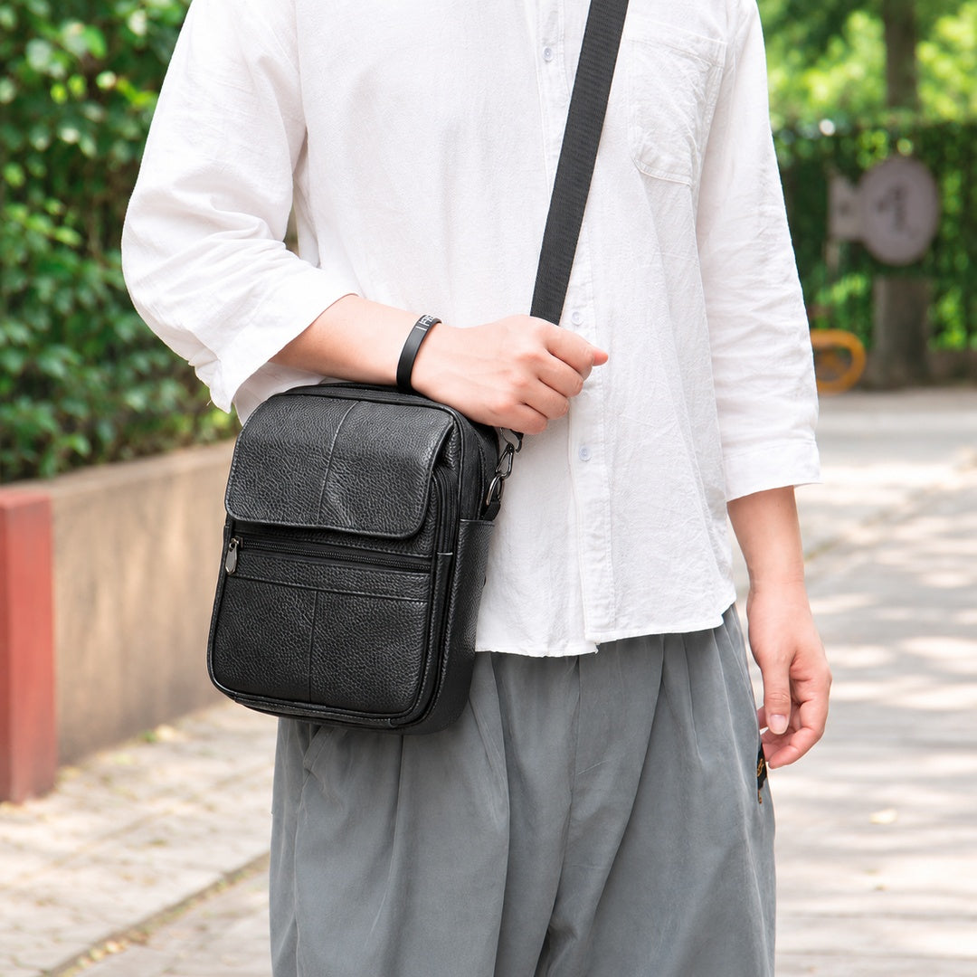A person wearing a white shirt and gray pants carries a black leather crossbody bag with a textured front pocket and a black strap, set against an outdoor background with green foliage.