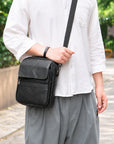 A person wearing a white shirt and gray pants carries a black leather crossbody bag with a textured front pocket and a black strap, set against an outdoor background with green foliage.