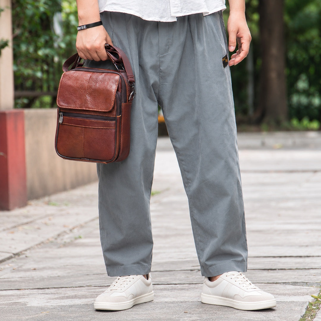 A person outdoors wearing loose gray pants and white sneakers, holding a brown leather crossbody bag. The background shows a blurred walkway and greenery.