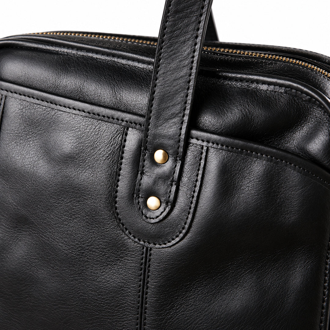A close-up of the black leather briefcase shows two brass-tone rivets securing a rounded handle tab over stitched reinforcement.