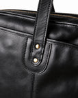 A close-up of the black leather briefcase shows two brass-tone rivets securing a rounded handle tab over stitched reinforcement.
