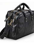 A black leather briefcase is shown at a side angle with twin zip compartments, leather zipper pulls and a brass-tone strap clasp.