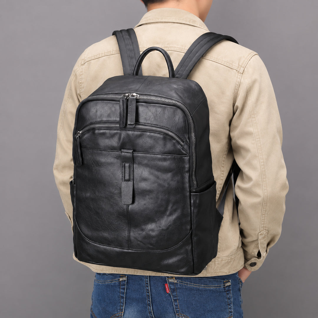 A man viewed from behind wearing a black leather backpack over a beige jacket and blue jeans, set against a solid gray studio background.