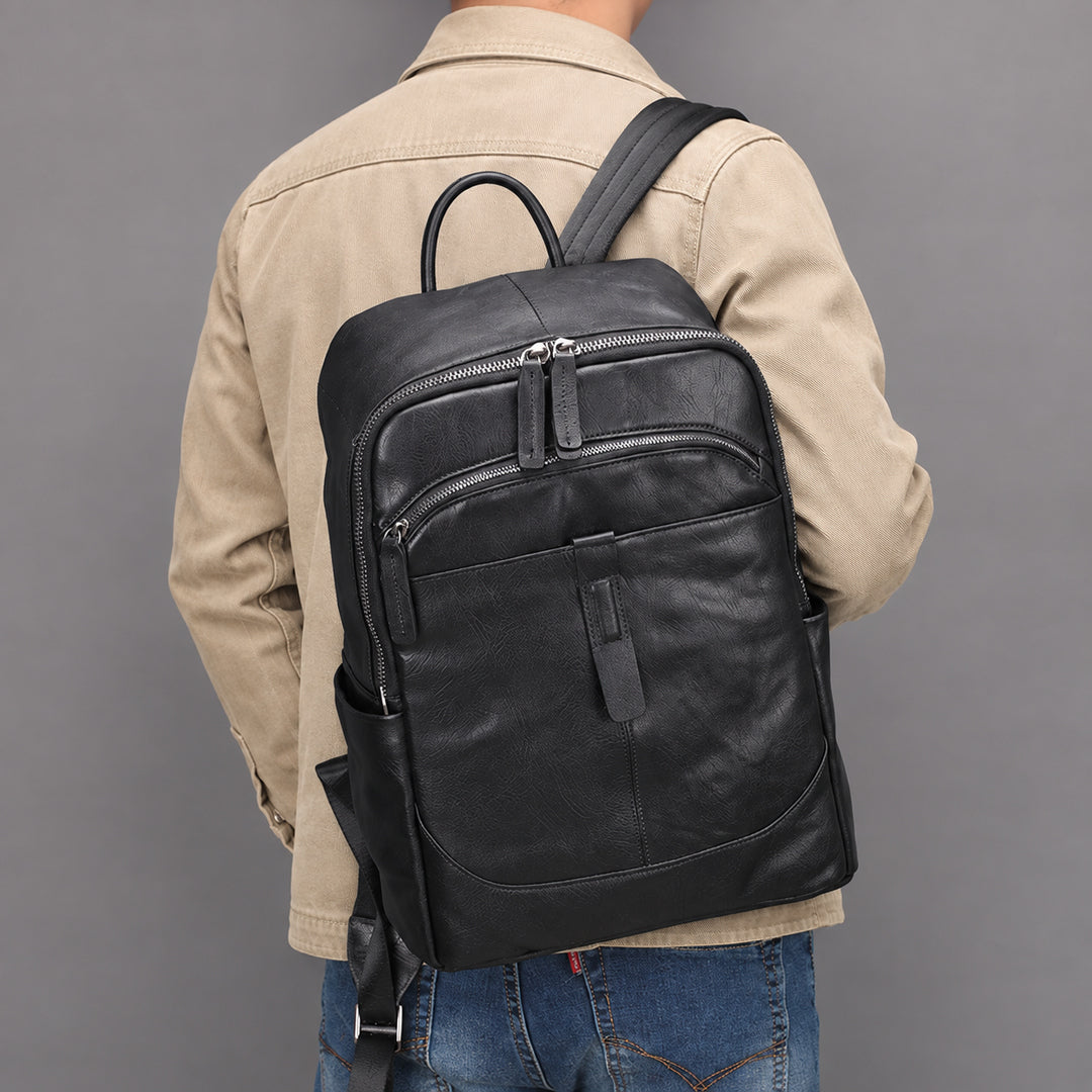 A man viewed from behind wearing a black leather backpack with multiple zippered compartments, a beige jacket, and blue jeans against a solid gray background.