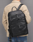 A man viewed from behind wearing a black leather backpack with multiple zippered compartments, a beige jacket, and blue jeans against a solid gray background.
