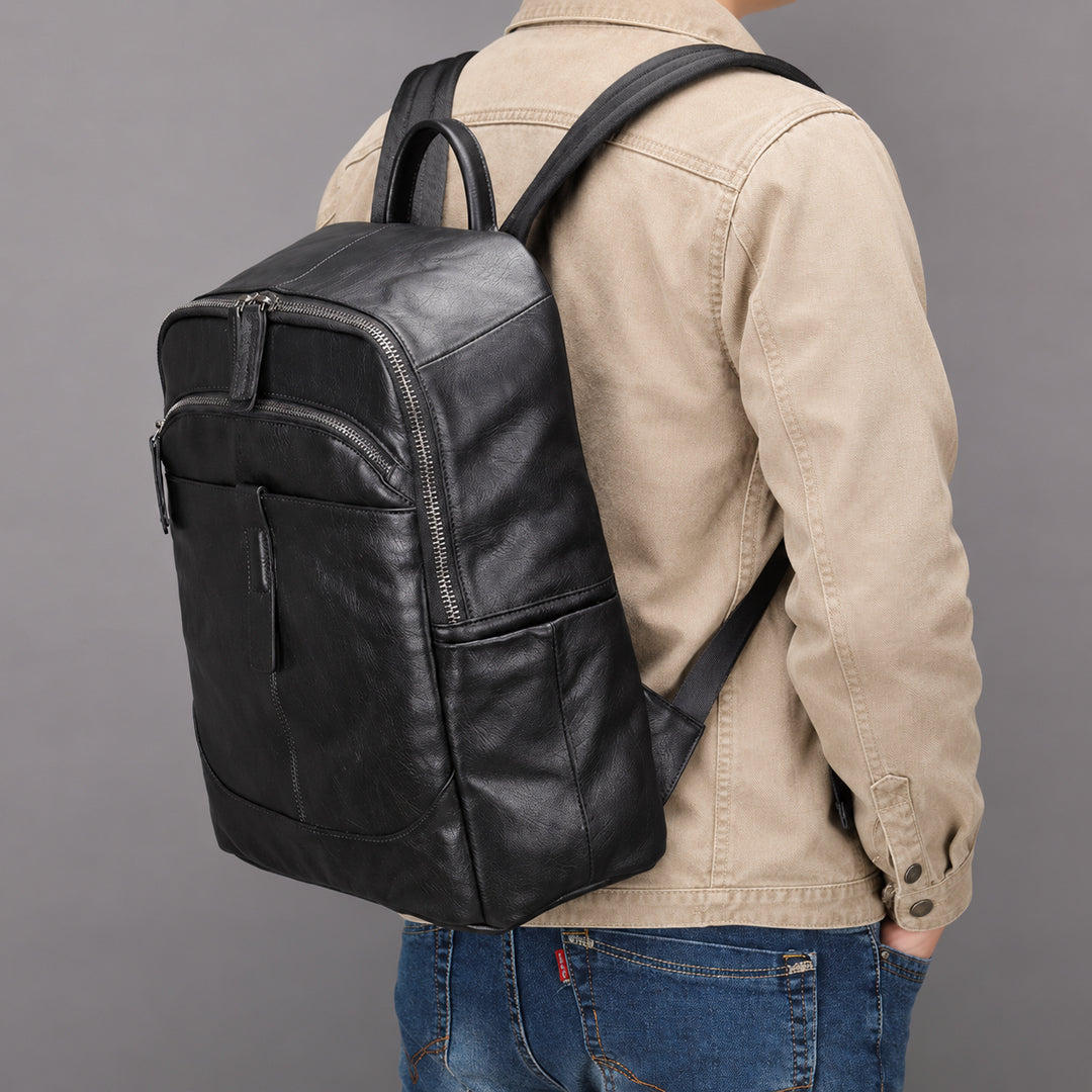 A black leather backpack with multiple zippered pockets, shown from the back on a model wearing a beige jacket and blue jeans against a solid gray background.