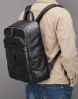 A black leather backpack with multiple zippered pockets, shown from the back on a model wearing a beige jacket and blue jeans against a solid gray background.