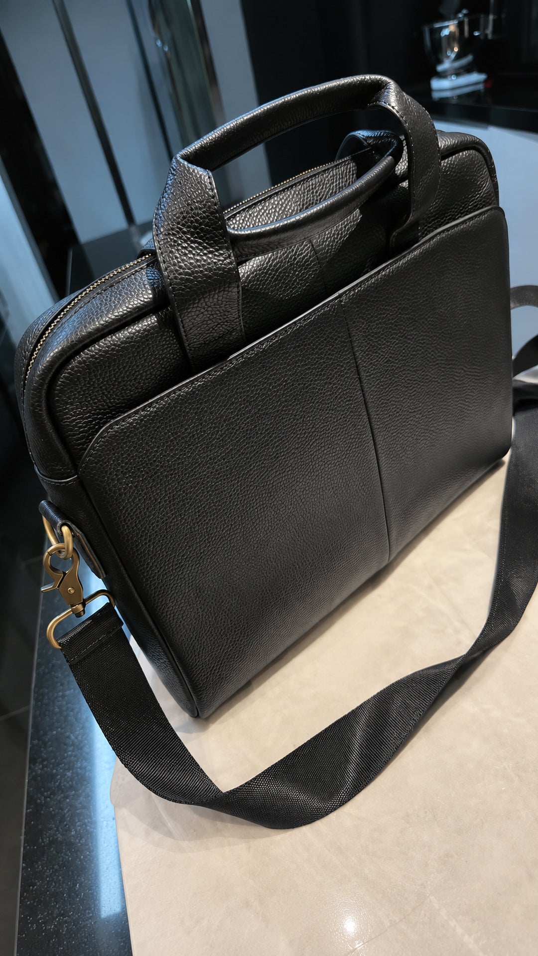 A black pebbled leather briefcase or laptop bag with dual top handles and a detachable shoulder strap, resting on a light beige surface in an indoor setting.